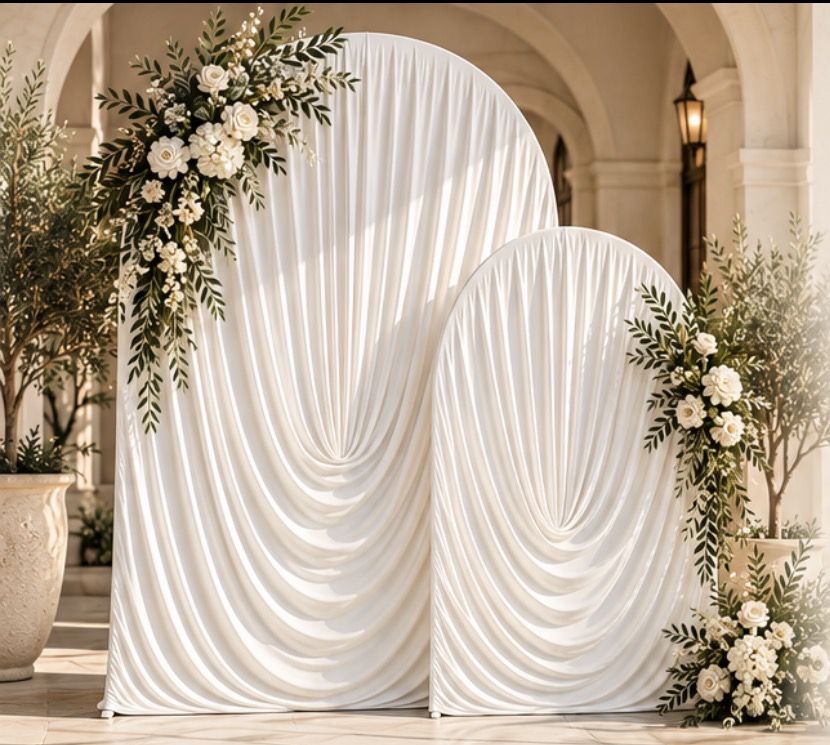Two ivory draped arch backdrops with white roses and olive greenery under a stone archway